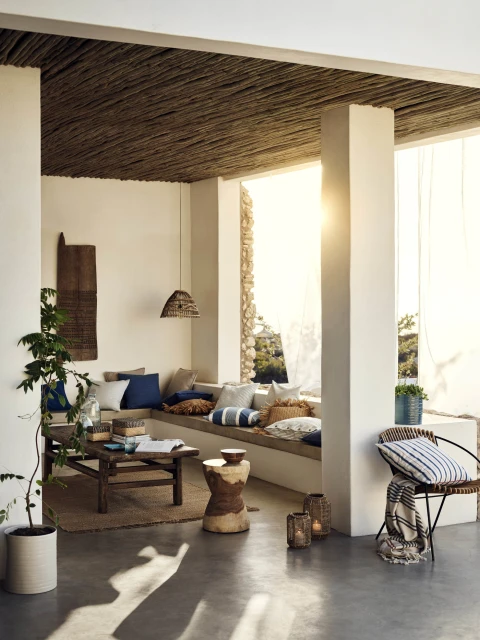 Sun-drenched terrace featuring built-in seating with pillows, rustic wood furniture, woven ceiling, and potted plants in a stylish outdoor living space.