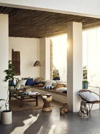 Sun-drenched terrace featuring built-in seating with pillows, rustic wood furniture, woven ceiling, and potted plants in a stylish outdoor living space.
