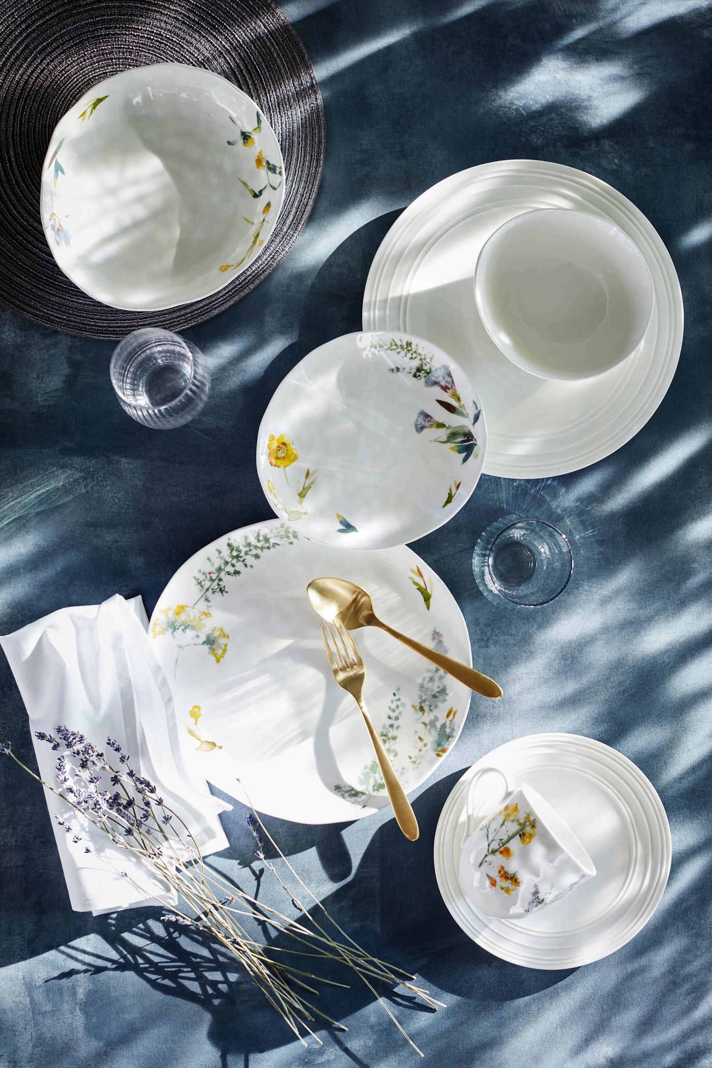 Overhead shot of an arrangement with floral-patterned plates, gold flatware, glasses, and a sprig of lavender on a blue tablecloth.