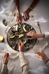 Overhead view of hands around a large tray of oysters on ice with champagne glasses and gold cutlery on a white tablecloth.