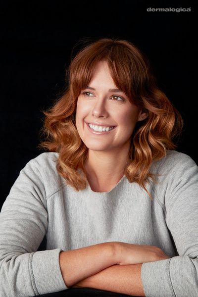 Portrait of a smiling adult woman with shoulder-length wavy red hair, wearing a gray long-sleeved shirt against a dark background.