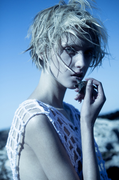 Portrait of a young woman with short blonde hair and white crocheted top, holding a rock fragment against a blue sky background.