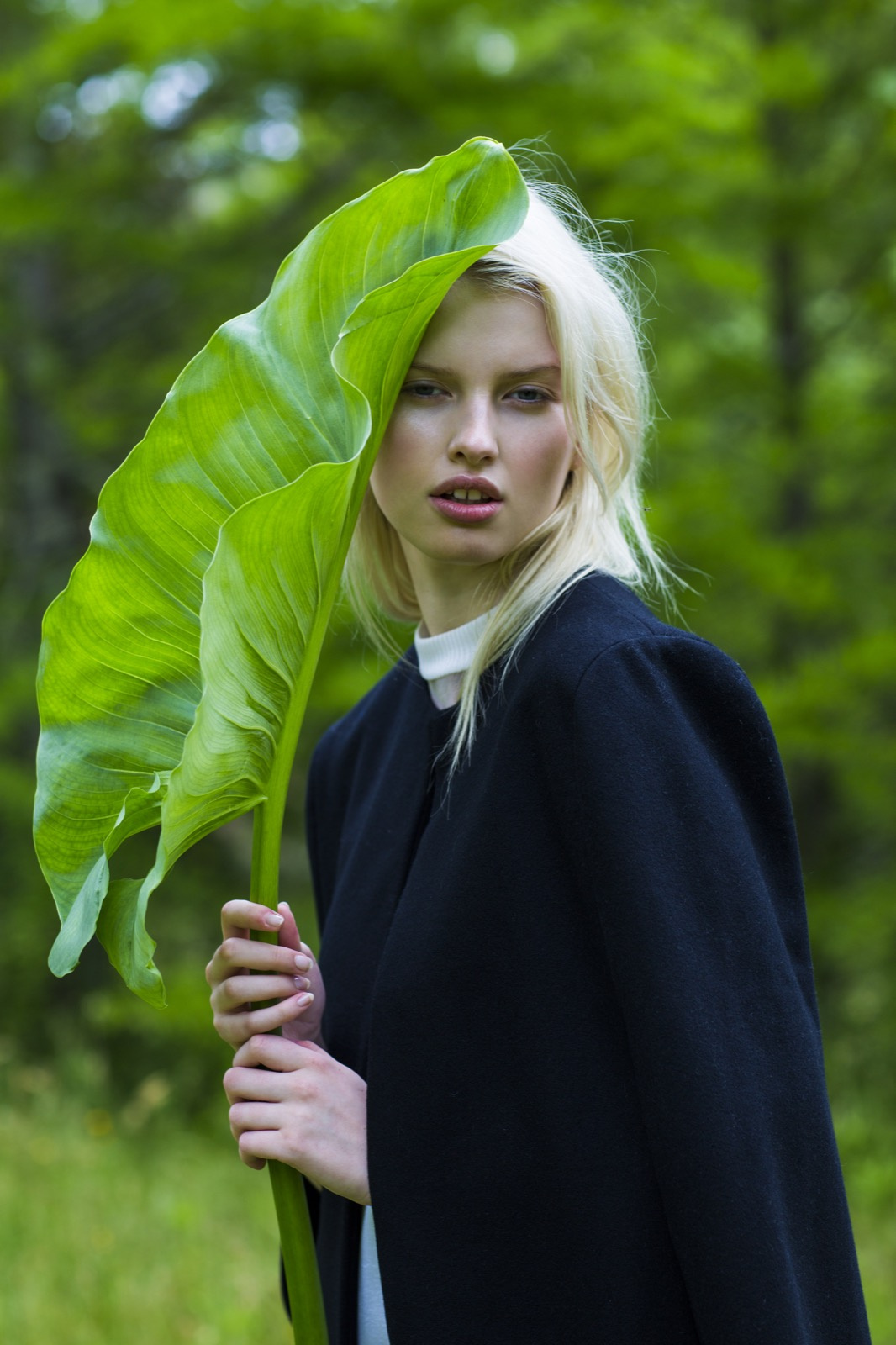 Young woman with blond hair and a black cape posing with a large green leaf in an outdoor forest setting, fashion portrait.