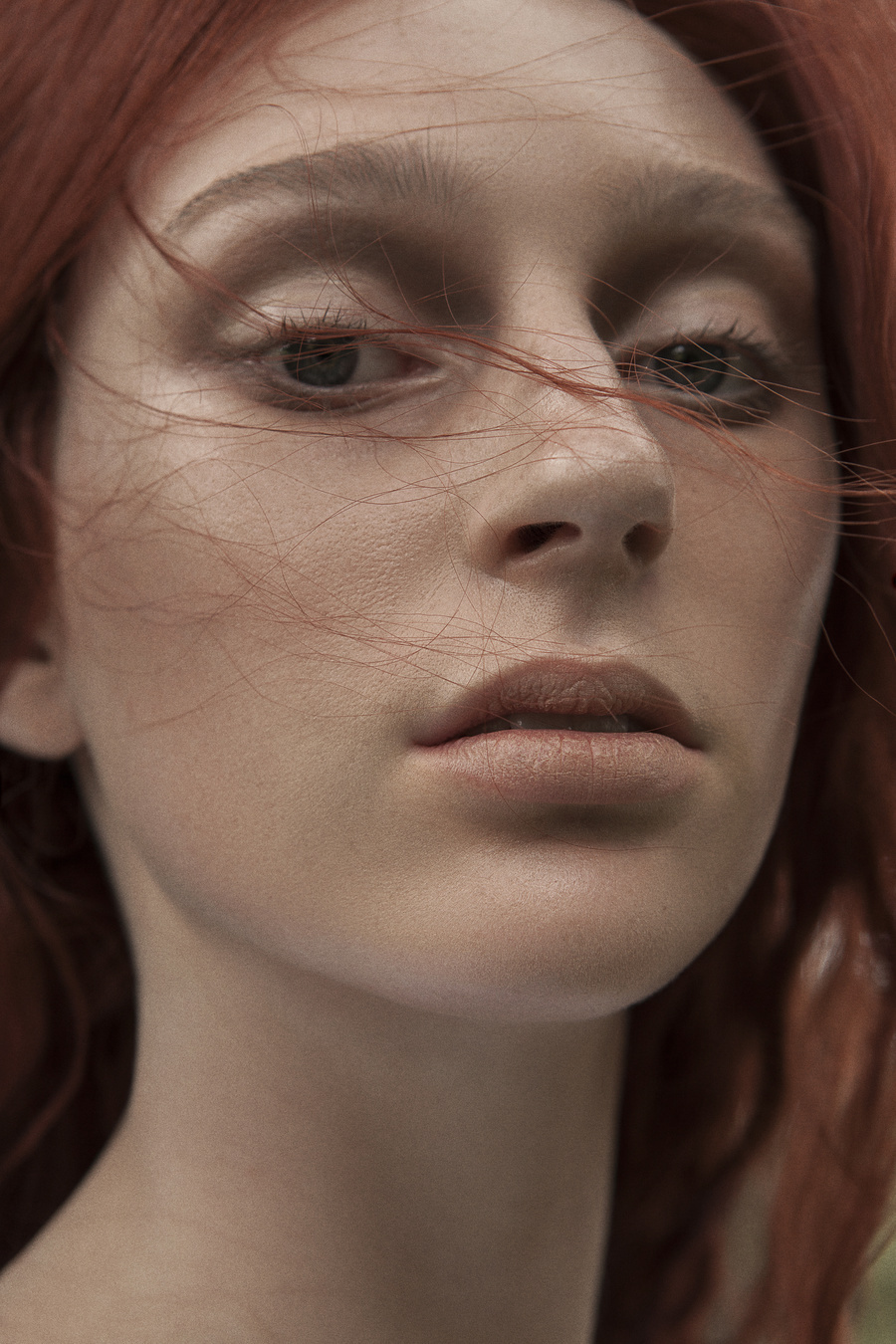 Close-up portrait of a woman with red hair looking directly at the camera, soft natural light, and a blurred green outdoor background.