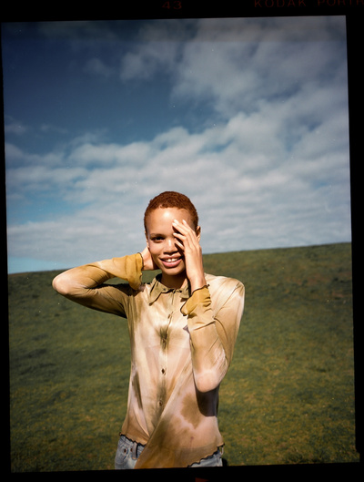 Woman with short hair posing in a field with a blue sky and clouds, wearing an ombre-colored shirt. Portrait shot.
