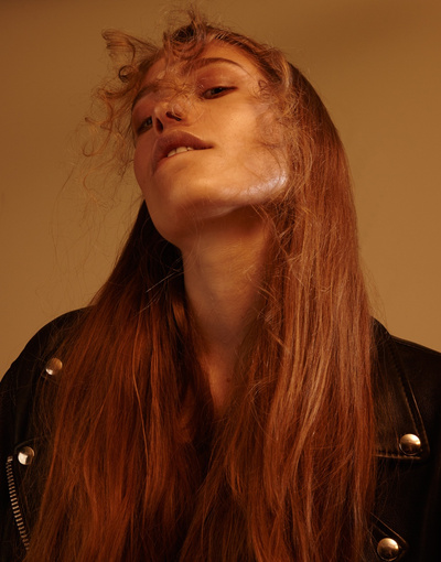 Portrait of a young adult woman with long red hair wearing a black leather jacket. Warm-toned indoor setting with soft lighting.