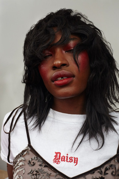 Close-up of an adult female with dark hair and red makeup, wearing a t-shirt and dress, against a plain white background.