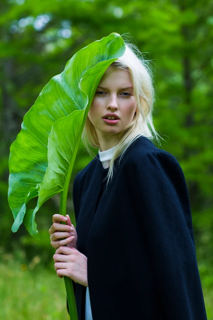 Blonde woman holding a large green leaf partially hiding her face outdoors in a forest.
