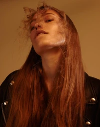 Low-angle portrait of a woman with long red hair wearing a black leather jacket against a warm amber background.