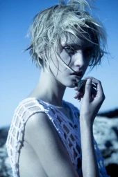 Close-up portrait of a woman with wet blonde hair wearing a white open-work crochet top against a bright blue sky.