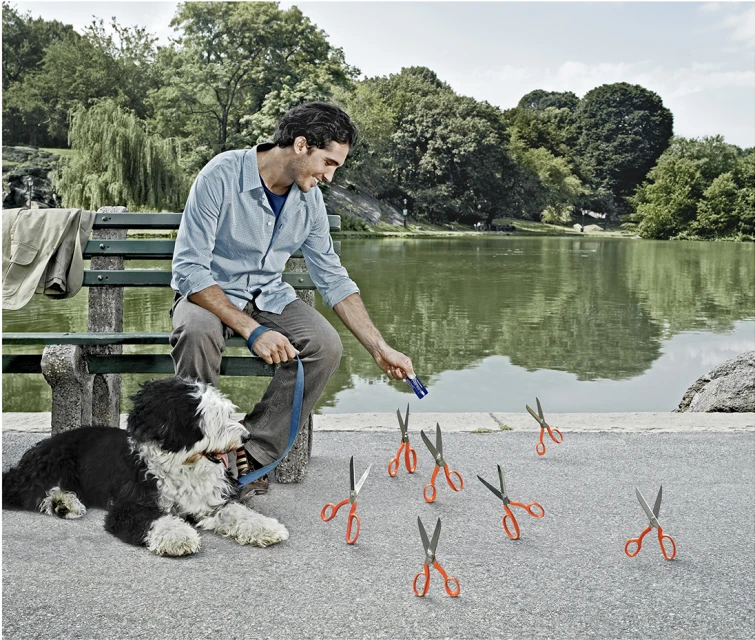 Man sitting on park bench with his dog, holding a treat with multiple scissors on the ground near a pond and trees.