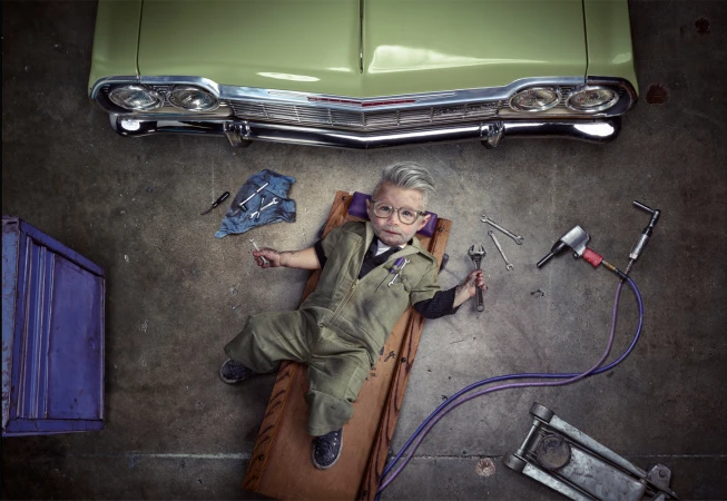 Child mechanic lying under a classic green car with wrenches, toolbox, and auto shop tools in a high-angle, vintage-style photograph.
