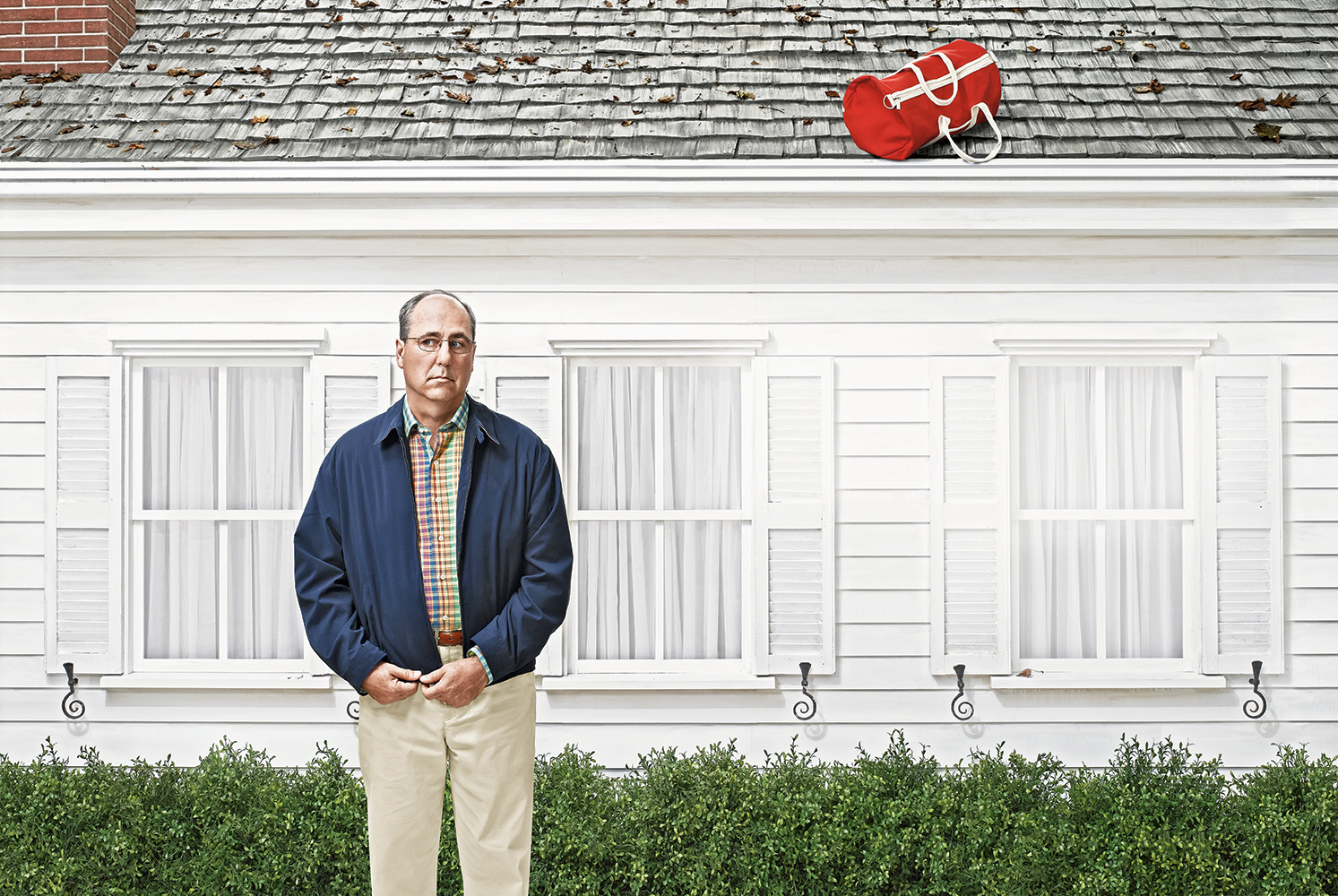 An adult man with a serious expression stands in front of a white house with a red duffel bag on the roof, creating a humorous scene.