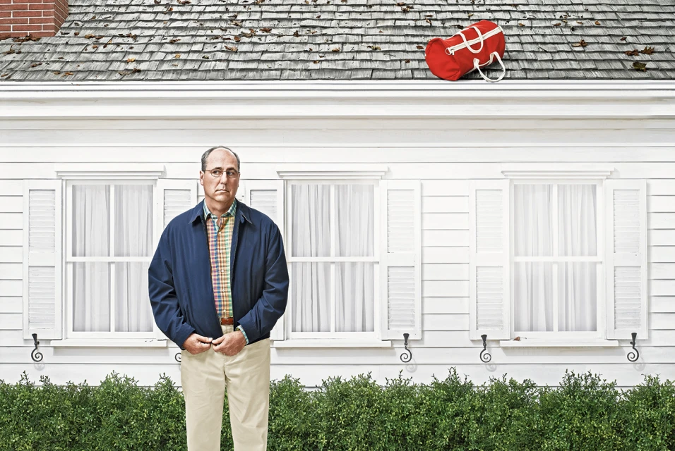 An adult man with a serious expression stands in front of a white house with a red duffel bag on the roof, creating a humorous scene.