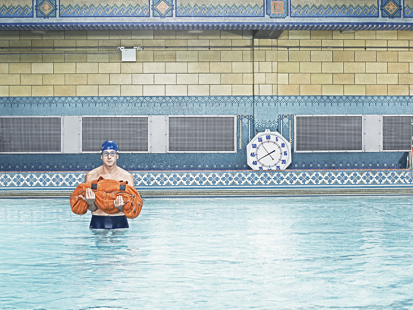 Man in swimming trunks and swim cap stands waist-deep in a pool holding an orange rescue device, with tiled walls and a clock behind him.