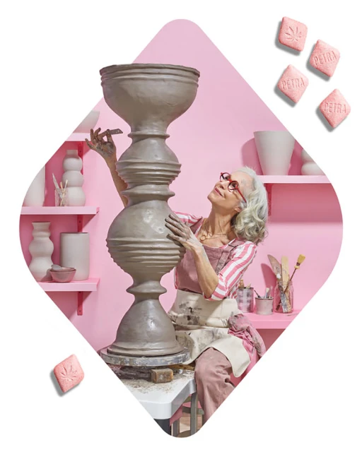 Older adult woman in glasses wearing an apron, sculpting clay vase in studio, with pink walls, lit by daylight, clay vase is the main subject