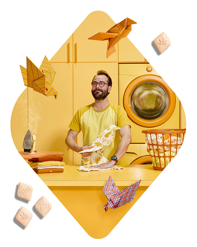 Bearded adult man folds a patterned textile on a yellow table with a vintage iron, folded clothes, origami birds, and washing machine.