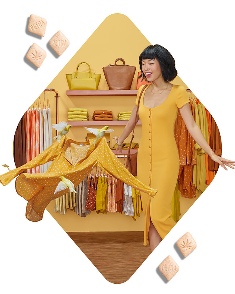 Smiling woman in yellow dress standing near rack of clothing with Petra edibles in foreground, fashion retail setting.