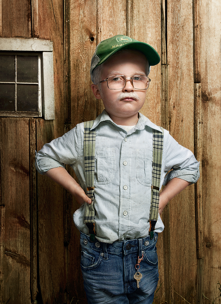 Child styled as an old man wearing glasses, suspenders, and a baseball cap standing in front of a rustic wooden wall.