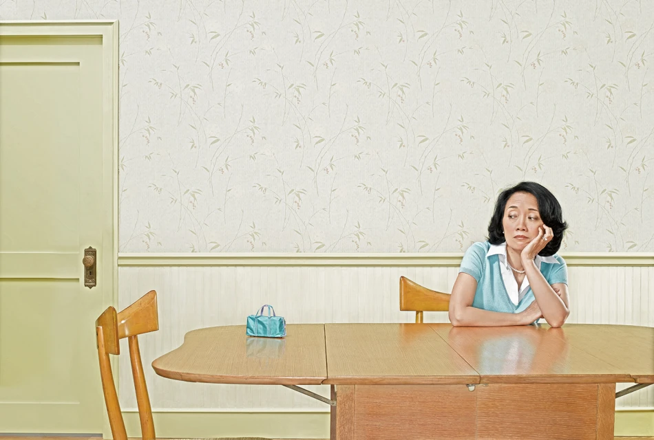 Asian woman sits at wooden table with teal purse, looking thoughtful in a pastel interior with patterned wallpaper and green door.