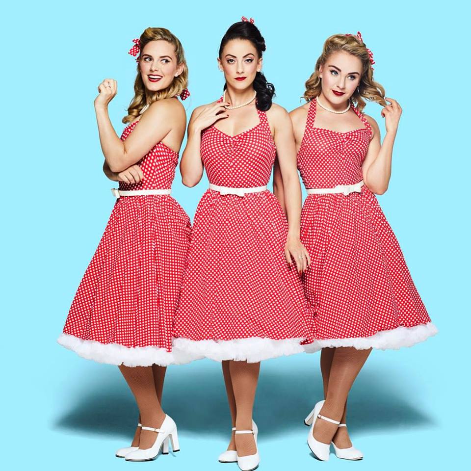 Three women in red polka dot dresses with white trim and white heels stand close together against a light blue backdrop, smiling.