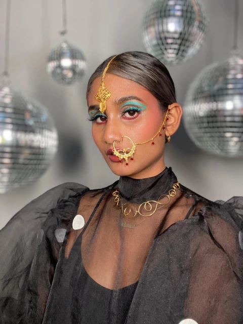Portrait of a young woman with gold jewellery, bright makeup, and sheer black top, posing in front of disco balls.