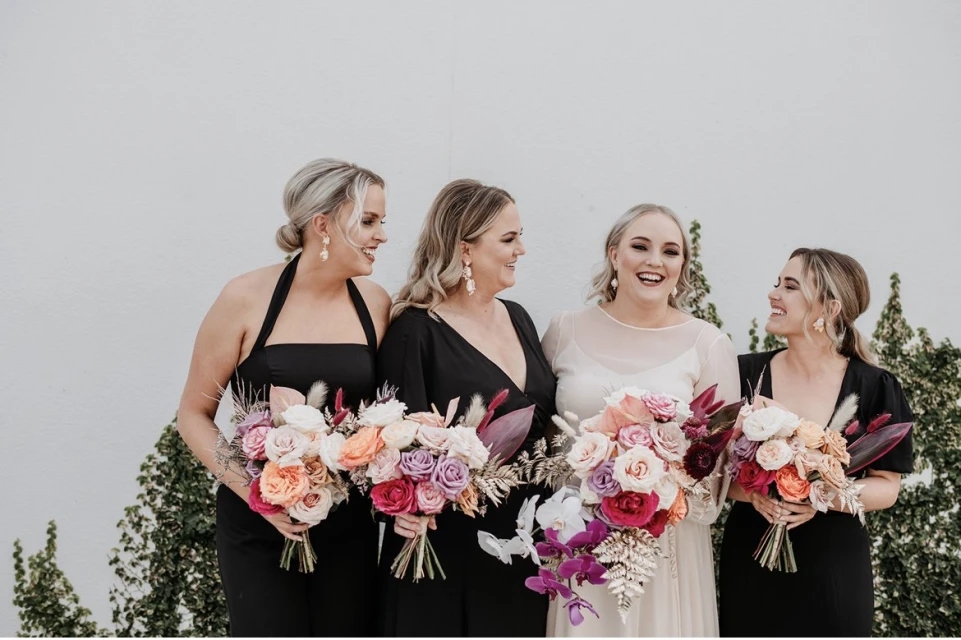 Four women in black dresses holding floral bouquets are smiling outside on a sunny day. Light grey background and green ivy. 