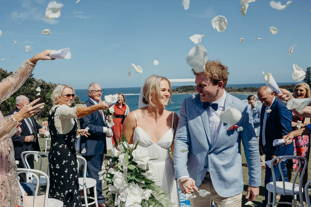 A bride and groom walk hand-in-hand as white flower petals fall around them, celebrating their outdoor wedding ceremony with guests.