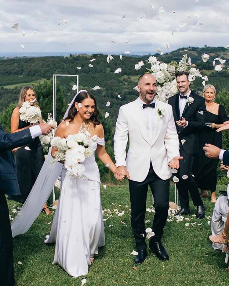 A joyous wedding scene featuring a bride and groom walking hand-in-hand amidst falling white flower petals on a grassy field.