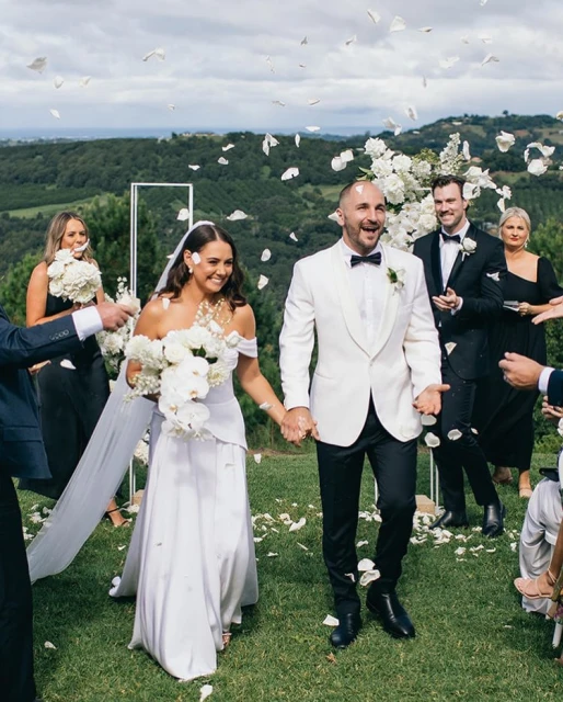 A joyous wedding scene featuring a bride and groom walking hand-in-hand amidst falling white flower petals on a grassy field.