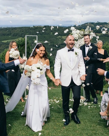 A joyous wedding scene featuring a bride and groom walking hand-in-hand amidst falling white flower petals on a grassy field.