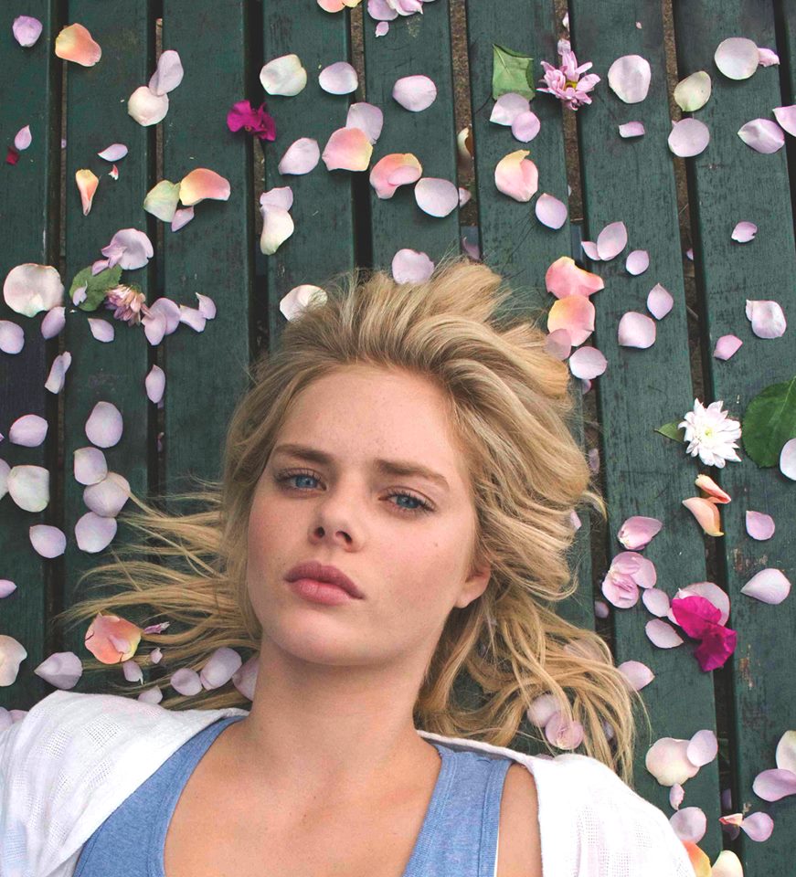 Blonde woman lays on a dark-green wooden surface covered with pink and white flower petals, looking up at the camera in a close-up shot.