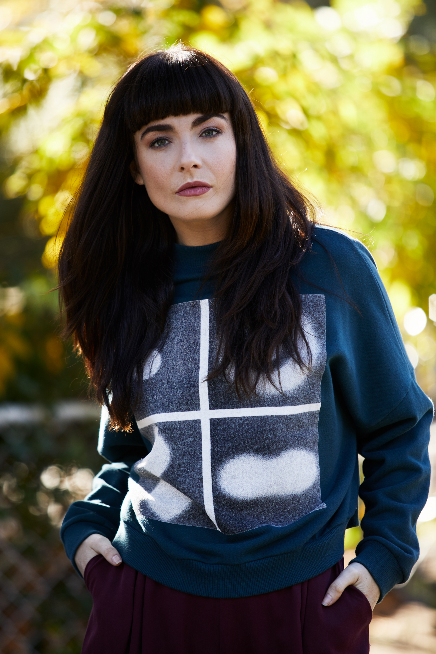 Portrait of a brunette woman with bangs wearing a teal sweater with a graphic design, standing outside with a blurred green and yellow background.