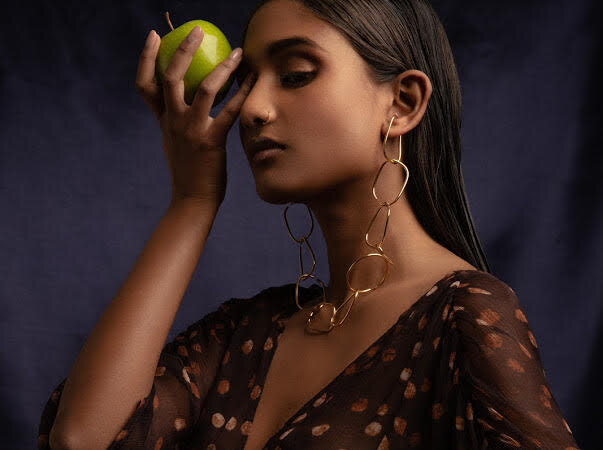 Woman poses in brown patterned dress with statement gold earrings, holding a green apple in front of a dark blue background.