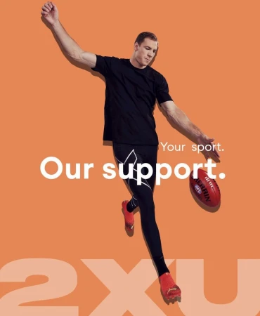 Athletic man in black activewear kicks an Australian rules football against an orange backdrop for a sports advertisement.