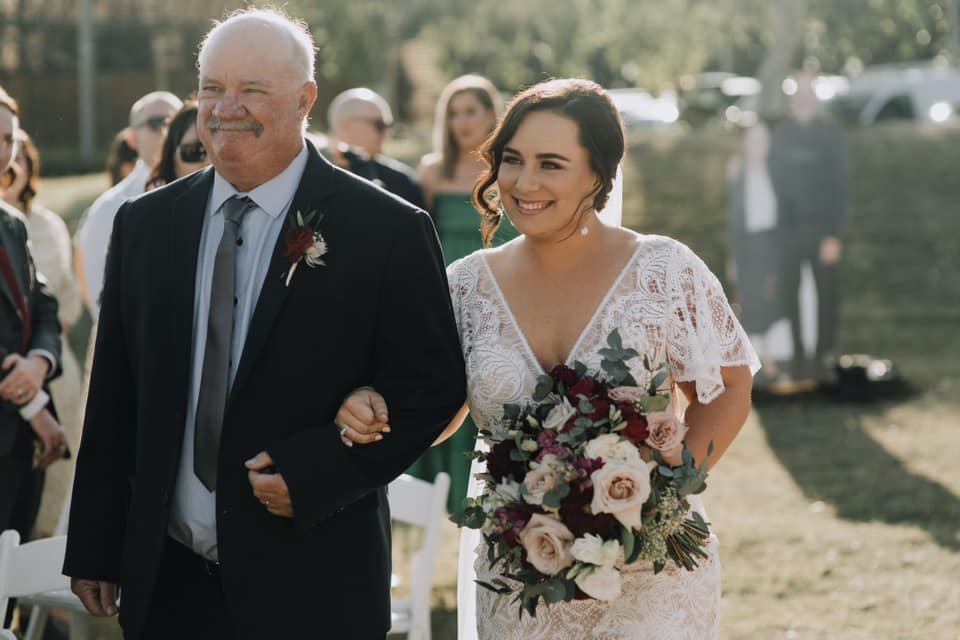 Bride and father walking outdoors on wedding day. The bride is holding a bouquet with pink roses, while the father wears a suit.