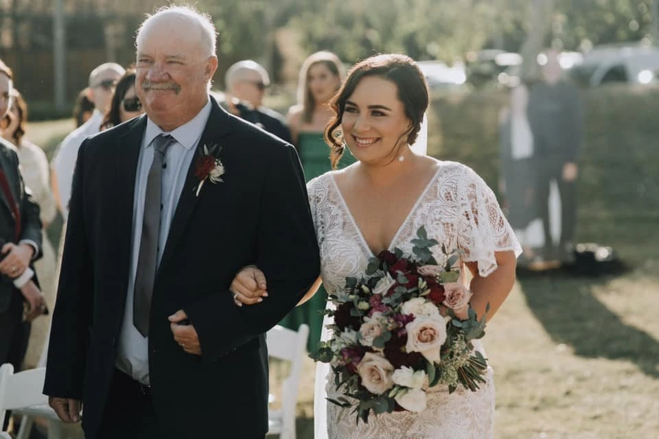 Bride and father walking outdoors on wedding day. The bride is holding a bouquet with pink roses, while the father wears a suit.