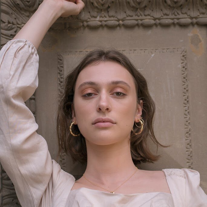 Close-up of a woman with hoop earrings posing against a decorative wall with an arm raised, in a portrait orientation.