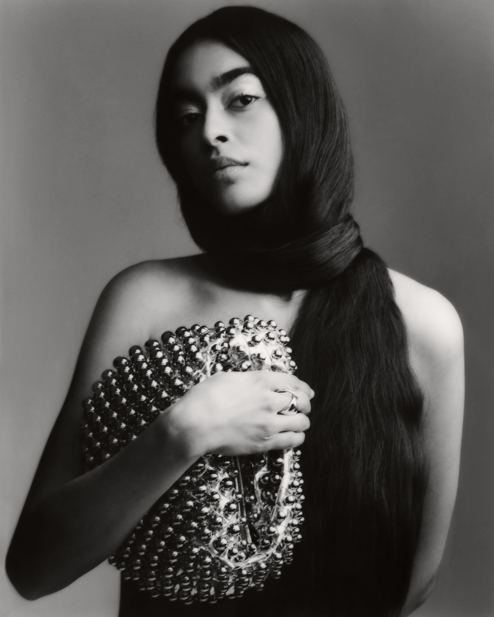 Portrait of a woman with long dark hair wrapped around her neck, holding a silver beaded bag, set against a grey background.