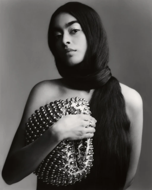Portrait of a woman with long dark hair wrapped around her neck, holding a silver beaded bag, set against a grey background.