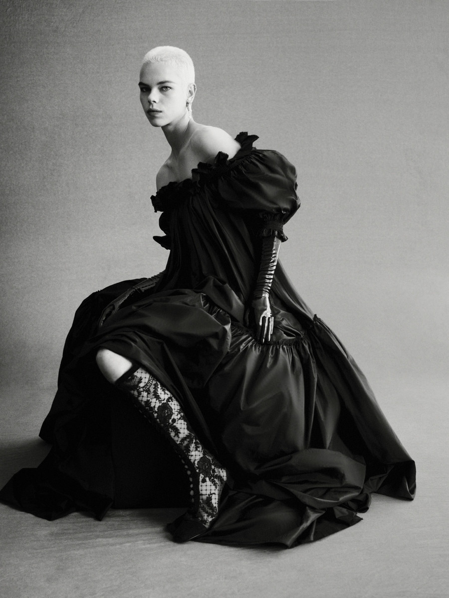 Monochrome portrait of a woman with short hair wearing a voluminous black dress, lace boots, and gloves, seated in a photo studio.