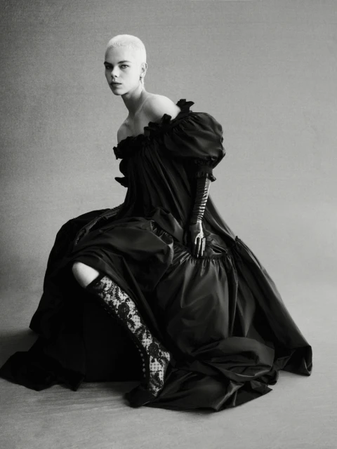 Monochrome portrait of a woman with short hair wearing a voluminous black dress, lace boots, and gloves, seated in a photo studio.