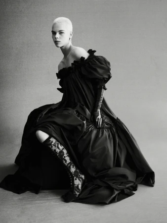 Monochrome portrait of a woman with short hair wearing a voluminous black dress, lace boots, and gloves, seated in a photo studio.