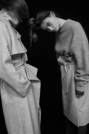 Monochrome fashion portrait featuring two women wearing belted coats and sweaters against a dark background.