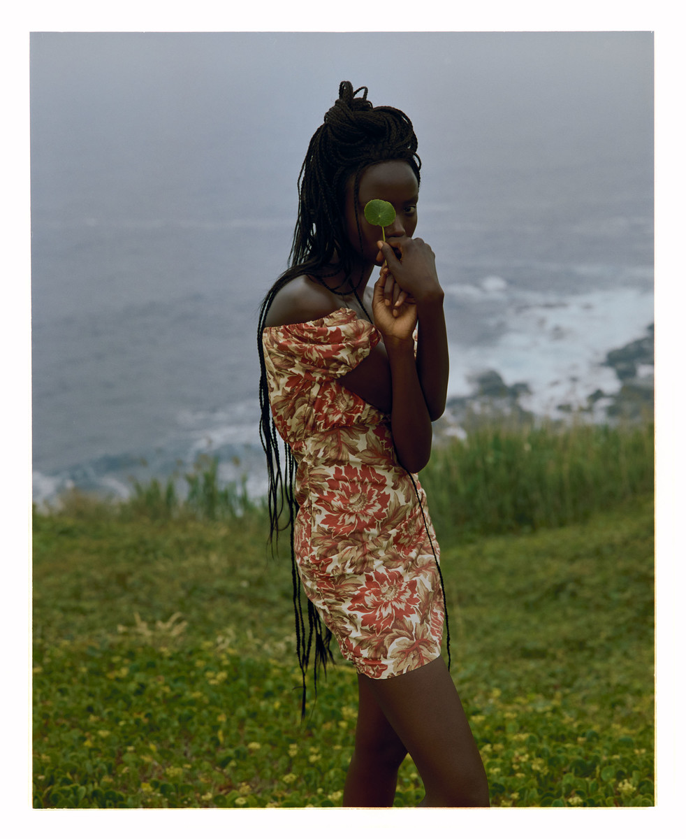 A dark-skinned woman stands outdoors by the ocean in a floral dress, holding a green leaf in front of her eye, with a cloudy sky.
