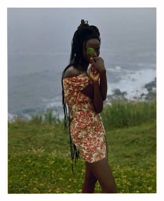 A dark-skinned woman stands outdoors by the ocean in a floral dress, holding a green leaf in front of her eye, with a cloudy sky.