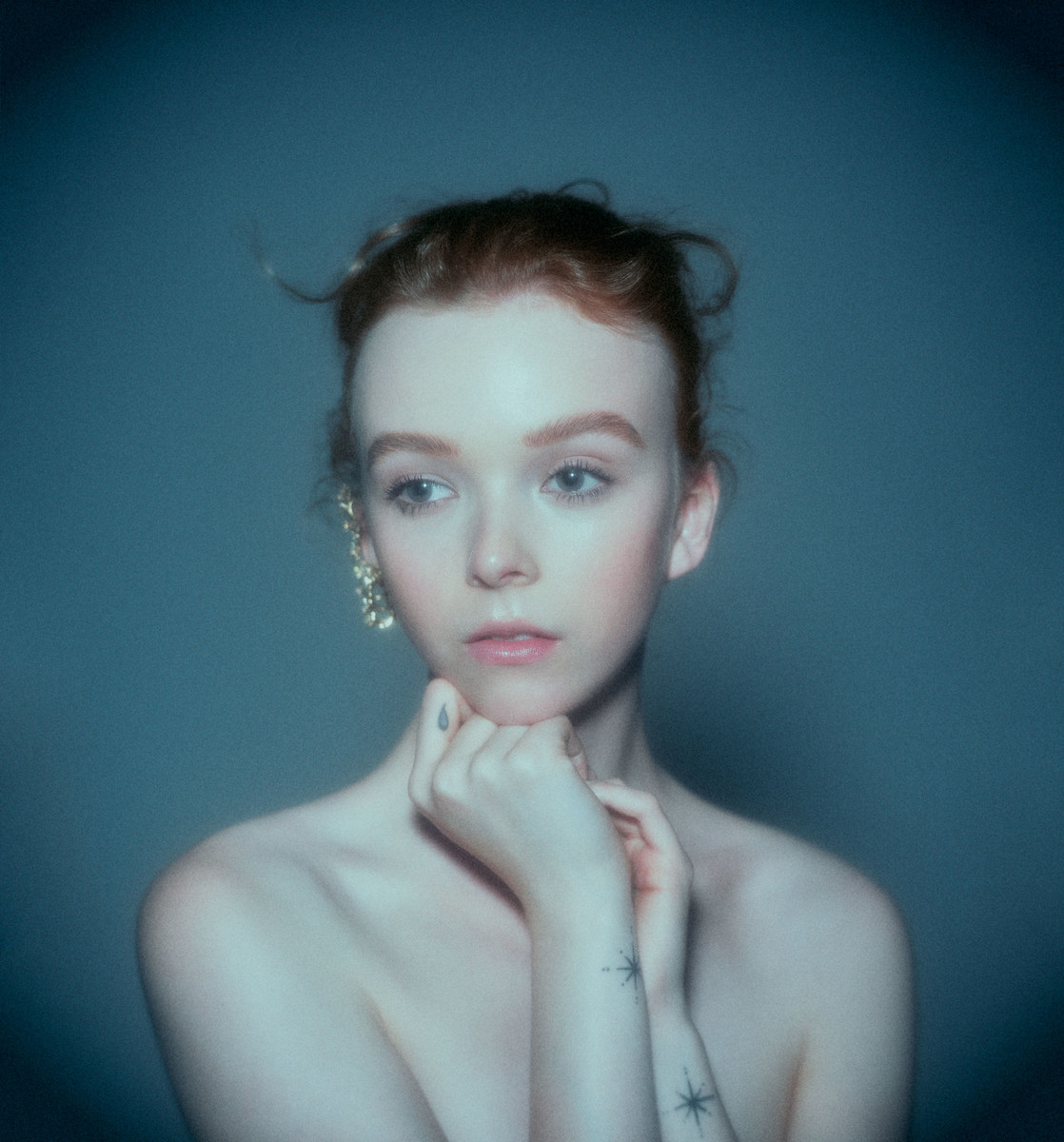 Portrait of a redheaded woman with hands under chin, wearing an ear cuff and small wrist tattoos against a dark teal background.