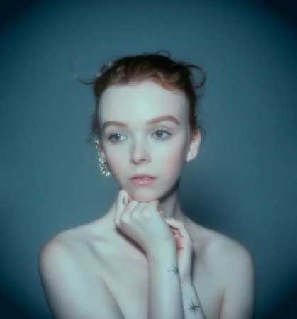 Portrait of a redheaded woman with hands under chin, wearing an ear cuff and small wrist tattoos against a dark teal background.