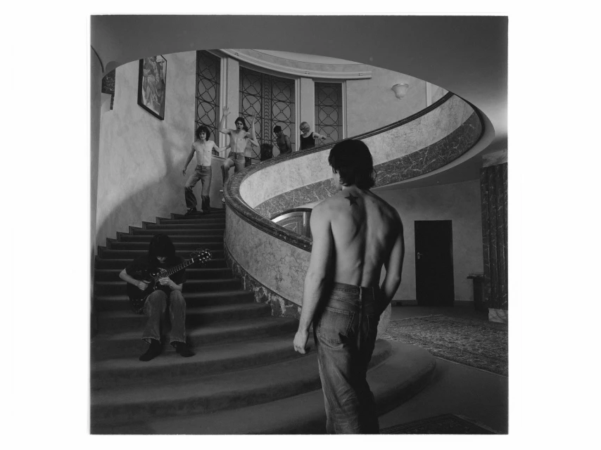 Black and white shot of shirtless man on stairs with guitar player and other people on a spiral staircase indoors with arched windows.