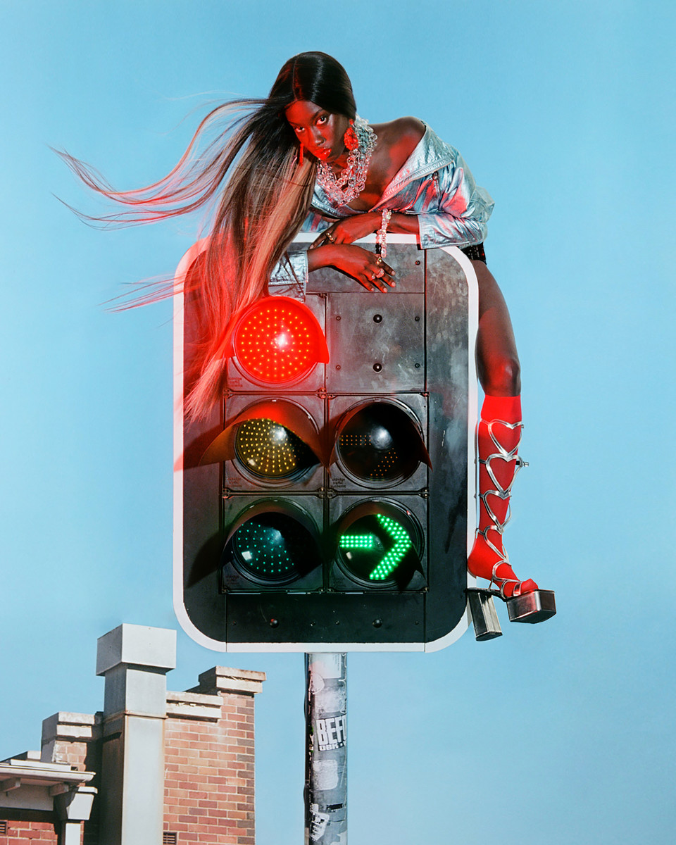 Fashionable black woman poses atop traffic signal in silver blouse, platform heels, and long hair against a blue sky backdrop.